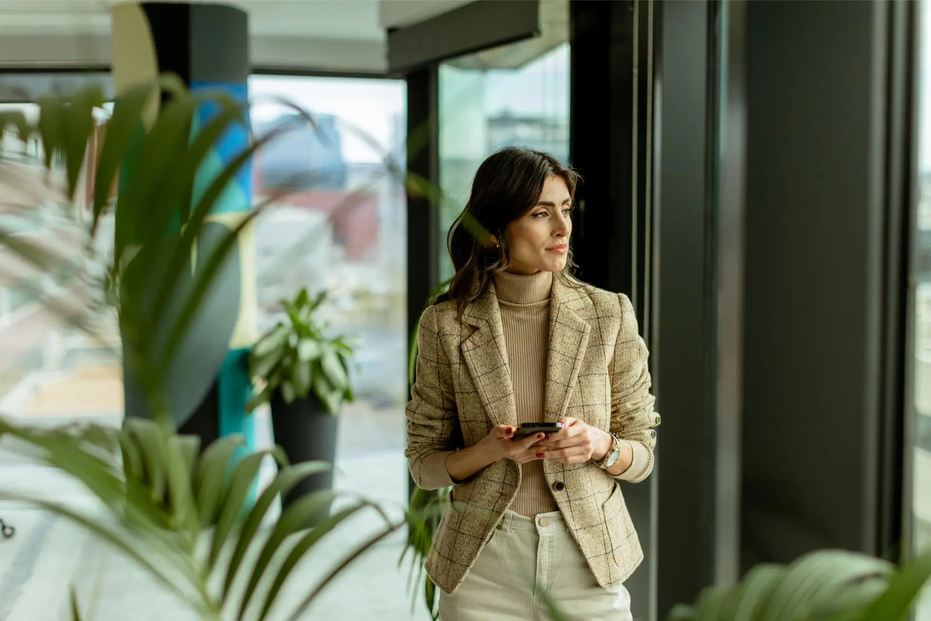 Stylish lady gazes out a window, holding a smartphone, amidst the urban allure