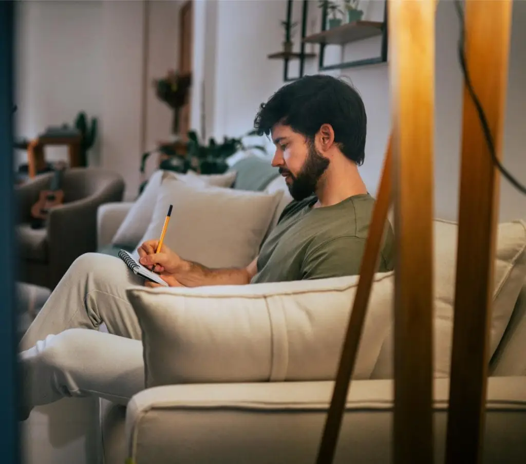 Window view of a Hispanic Brazilian beautiful brunette man relaxing in his living room sitting on a light colored couch taking notes on notepad with a white wall metal and wood shelves plants an armchair pillows and a throw in the background wearing a green t-shirt and white pants leaning on a pillow