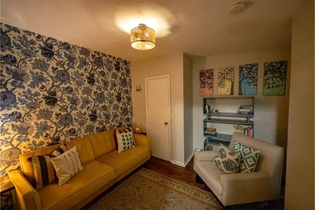 Warm therapy space with a yellow couch, floral wallpaper, and a bookshelf.